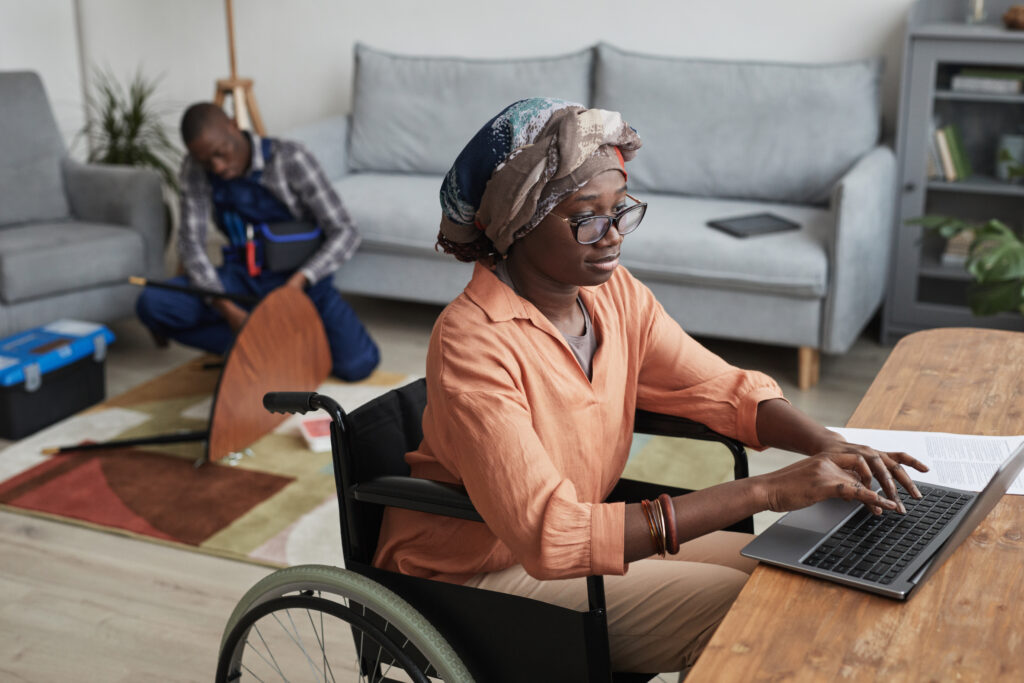 Portrait of disabled woman using wheelchair and working from home with a handyman assembling furniture in background.